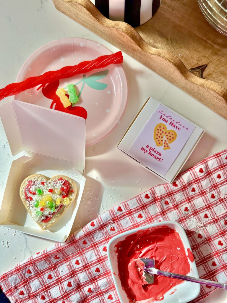 Heart shaped cookie decorated to look like a pizza topped with shredded white chocolate and candy and placed in mini pizza boxes to look like mini pizza heart cookies