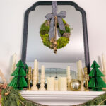 Mantle decorated for Christmas with candles and accordian paper Christmas trees. A mirror is above mantle with black frame. In the middle of the mirror is a wreath made of moss hanging by a black and white striped ribbon with bells hanging.